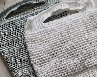 May include: Two handmade crochet tote bags. One is a light gray color, and the other is white. Both bags have a rectangular shape with a handle opening at the top. The crochet stitch creates a textured, open weave pattern.