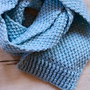 May include: A light blue knitted scarf featuring a textured stitch pattern throughout the main body and a ribbed border at the end. The scarf is crafted from yarn and displays a consistent, soft blue color across its entire length.