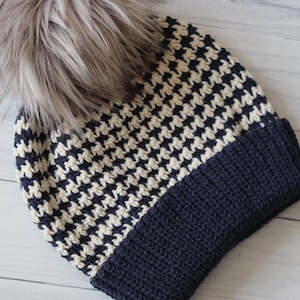 May include: A black and white houndstooth patterned beanie with a navy blue brim and a fluffy brown pom pom.