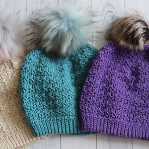 May include: Three crocheted hats with faux fur pom poms. The hats are beige, teal, and purple.