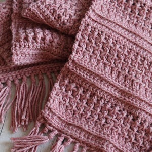 Puede incluir: Una bufanda de ganchillo rosa polvoriento con un patrón de puntadas texturizadas y flecos. La bufanda está doblada, mostrando el intrincado diseño y el color suave. El accesorio hecho a mano es acogedor.
