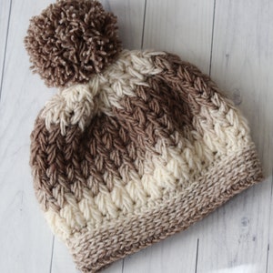 May include: A brown and white knitted beanie with a brown pom pom on top. The beanie has a cable knit pattern.