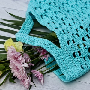 May include: A turquoise crocheted tote bag with a wide, open top. The bag is filled with a bouquet of pink and yellow flowers.
