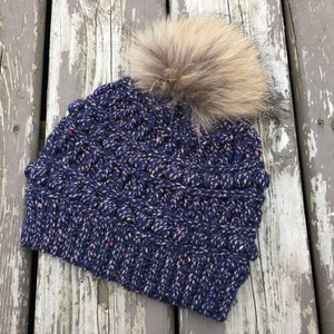 May include: A navy blue knit beanie with a white and pink fleck pattern and a brown faux fur pom pom.