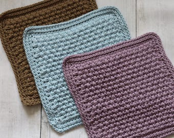 May include: Three crocheted square washcloths in shades of brown, blue, and purple. Each cloth has a textured pattern and a finished border. The washcloths are arranged on a light wood surface.