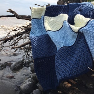 May include: A blue, white, and light blue crocheted blanket with a textured pattern. The blanket is draped over a driftwood branch on a beach.