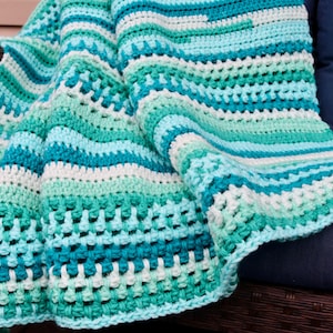 May include: A crocheted blanket with stripes of teal, turquoise, mint green, and white. The blanket is folded and draped over a wicker chair.