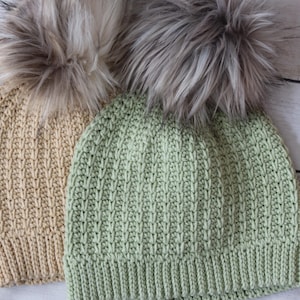 Puede incluir: Dos gorros de punto con pompones de piel sintética. Uno es de color beige claro, el otro verde claro. Los gorros tienen un diseño de punto acanalado y se muestran sobre una superficie clara con vetas de madera. Los pompones son grises claros y oscuros.
