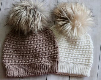 May include: Two knitted beanies with faux fur pom-poms. One is light brown, the other white. The beanies have a textured stitch pattern. They are displayed on a light wood surface.