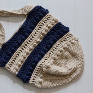May include: A crocheted tote bag with a beige and navy blue striped design. The bag has a wide, flat bottom and a single strap. The bag is decorated with small, round pom-poms.