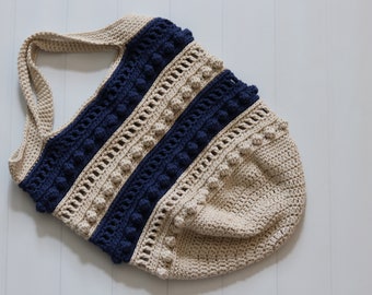 May include: A crocheted tote bag with a beige and navy blue striped design. The bag has a wide, flat bottom and a single strap. The bag is decorated with small, round pom-poms.
