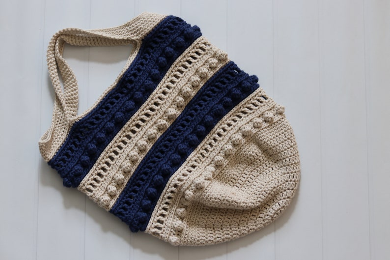 May include: A crocheted tote bag with a beige and navy blue striped design. The bag has a wide, flat bottom and a single strap. The bag is decorated with small, round pom-poms.