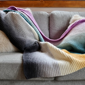 Peut inclure: Un plaid tricoté avec une texture côtelée et un motif dégradé multicolore passant par des nuances de gris foncé, gris clair, crème, jaune, bleu sarcelle et violet, drapé sur un canapé gris avec deux coussins en fausse fourrure.