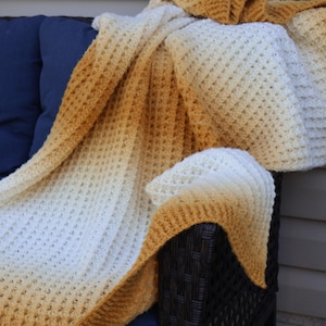 May include: A cozy, hand-knitted blanket in a waffle stitch pattern. The blanket transitions from a creamy white to a warm, golden-brown hue. It is draped over a dark blue cushion and a wicker chair, creating a comfortable and inviting aesthetic.