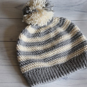 May include: A knitted winter hat with a gray and cream striped pattern. The hat has a gray brim and a cream-colored pom-pom on top. The hat is made of a soft, warm material, perfect for cold weather.