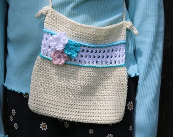 Pode incluir: Uma bolsa de crochê de cor creme com um design de flores de crochê azul e branco. A bolsa tem uma alça longa e é usada no ombro.
