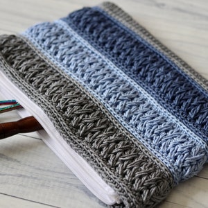 May include: A crocheted zippered pouch with a grey and blue colour gradient. The pouch is made with a textured stitch and has a white zipper. The pouch is open and has a pair of scissors and a wooden knitting needle inside.