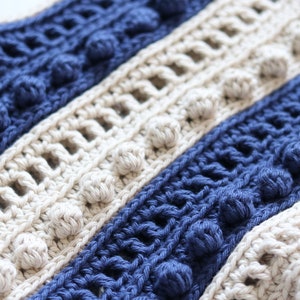 May include: Close-up of a crocheted fabric with alternating stripes of beige and navy blue. The fabric has a textured, bumpy surface with small, round, crocheted balls.