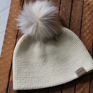May include: A white crocheted beanie with a fluffy white and brown pom pom on top. The beanie has a small brown leather label with the text "Nori Textures" on it.