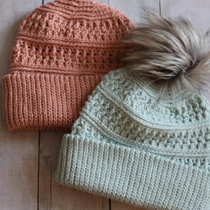 May include: Two knitted beanies in the image. One is a light coral color, and the other is a light blue-green color with a faux fur pom-pom. Both beanies have a ribbed brim and a textured stitch pattern on the crown.