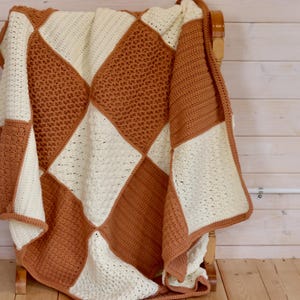 May include: A handmade crochet blanket with a geometric diamond pattern in cream and rust-orange. The blanket is draped over a wooden chair, showcasing the texture and color variations. The blanket is a cozy home decor item.