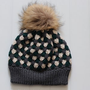 Puede incluir: Un gorro de punto de color gris, verde y crema con un pompón de piel sintética marrón.