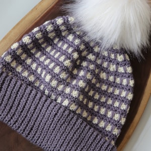 May include: A purple and cream crocheted beanie with a white faux fur pom pom. The beanie has a ribbed brim and a textured pattern.
