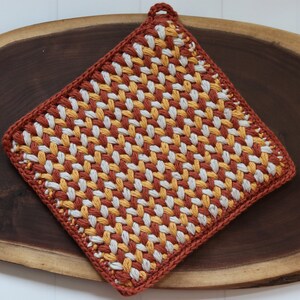 May include: A crocheted pot holder with a woven pattern in shades of orange, yellow, and white. The pot holder is lying on a wooden surface.