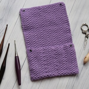 May include: A purple crocheted needle case with two pockets and four metal poppers. The case is lying on a white wooden surface with three crochet needles and a pair of scissors.