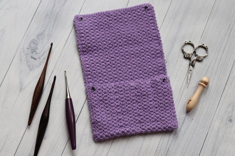 May include: A purple crocheted needle case with two pockets and four metal poppers. The case is lying on a white wooden surface with three crochet needles and a pair of scissors.