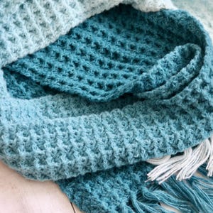 May include: A close-up of a handmade, textured scarf in shades of blue and teal. The scarf has a waffle-like pattern and is finished with white fringe. The soft, knitted material is folded over itself.