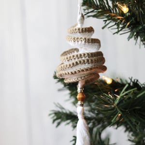 May include: A white and brown crocheted spiral Christmas tree ornament with a wooden bead and a white tassel hanging from a green artificial Christmas tree.
