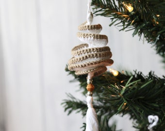 May include: A white and brown crocheted spiral Christmas tree ornament with a wooden bead and a white tassel hanging from a green artificial Christmas tree.