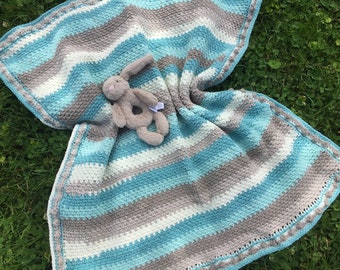 May include: A crocheted baby blanket with a striped pattern in shades of blue, gray, and white. The blanket has a crocheted border with small white pom-poms. A gray plush bunny toy is lying on the blanket.