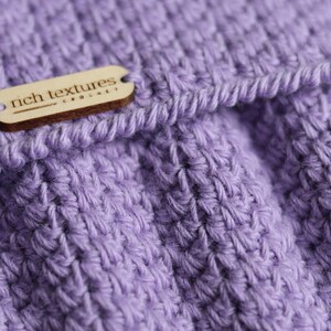 May include: A close-up of a purple crocheted fabric with a wooden tag that reads "rich textures crochet".