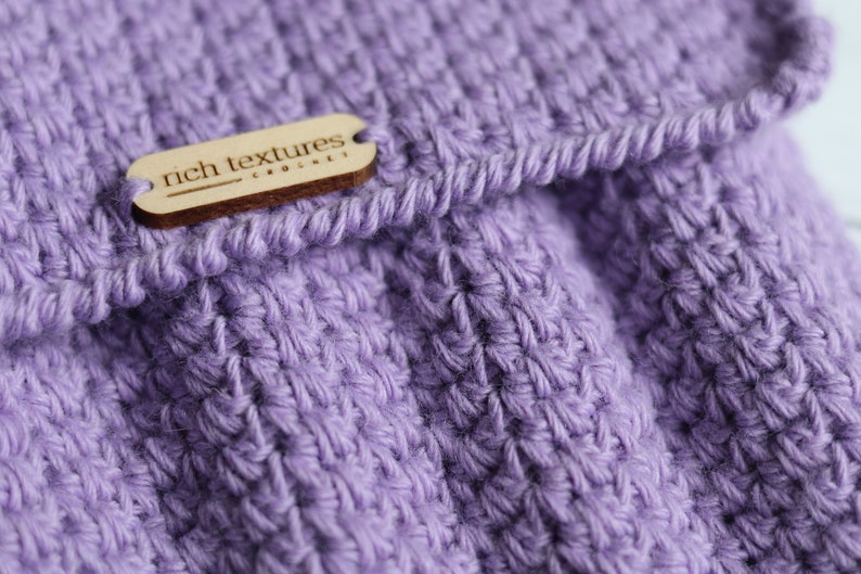 May include: A close-up of a purple crocheted fabric with a wooden tag that reads "rich textures crochet".