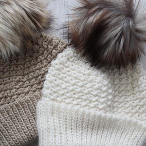 May include: Two knitted winter hats with faux fur pom poms. One hat is beige and the other is white.