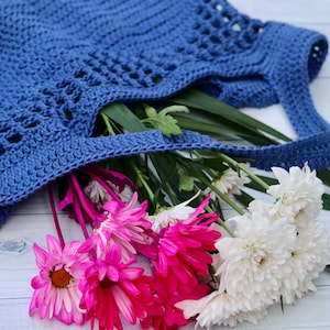 May include: A blue crocheted tote bag with a white and pink flower bouquet inside. The bag has a woven design and is lying on a white wooden surface.