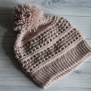 May include: A light pink crocheted beanie with a fluffy pom pom on top. The beanie has a textured stitch pattern and a ribbed brim.