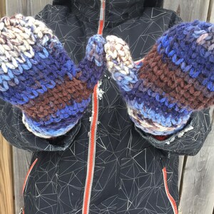 Könnte beinhalten: Ein Paar gestrickte Fäustlinge in Blau, Braun und Weiß. Die Fäustlinge haben ein gestreiftes Muster und sind aus einem dicken Garn gefertigt.