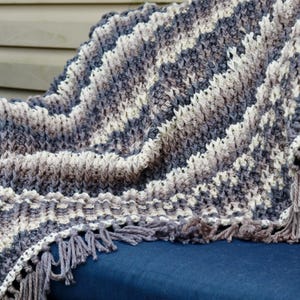 May include: A crocheted throw blanket with a striped pattern in shades of gray, white, and brown. The blanket has fringe along the edges.