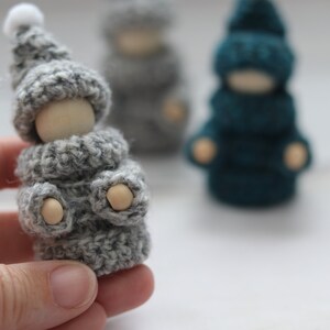 Op de afbeelding: Een grijze gehaakte gnome met een witte pomponhoed en een houten hoofd. De gnome draagt een grijze gehaakte trui en heeft houten handen.