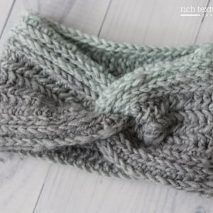 May include: A hand-knitted headband in a two-tone design. The lower section is a medium gray, transitioning to a light gray-green at the top. The headband features a twisted knot detail at the center, showcasing the textured knit.