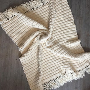May include: A cream-coloured crocheted throw blanket with a fringe border. The blanket is folded and draped on a wooden surface.