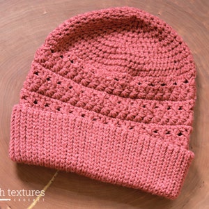 Peut inclure: Un bonnet en crochet de couleur rose poussiéreux avec un motif de point texturé. Le bonnet a un bord plié.