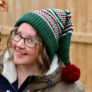 May include: A green, white, and red striped knit hat with a long pointed top and a red pom-pom. The hat is worn by a person with glasses and a brown jacket.