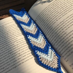 May include: A blue, white, and gray crocheted arrow bookmark. The bookmark is laying on top of an open book.