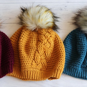 Op de afbeelding: Drie gebreide mutsen met nep bont pompoenen. De mutsen zijn in bordeauxrood, mosterdgeel en teal.