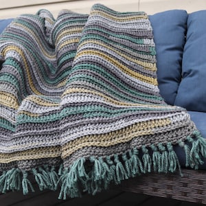 May include: A striped crocheted throw blanket with a fringe border in shades of grey, green, and yellow. The blanket is draped over a blue sofa.