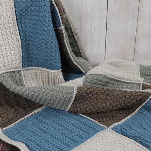 Op de afbeelding: Een gehaakt deken met een patchwork ontwerp in tinten bruin, blauw en beige. Het deken is gevouwen en gedrapeerd over een houten oppervlak.